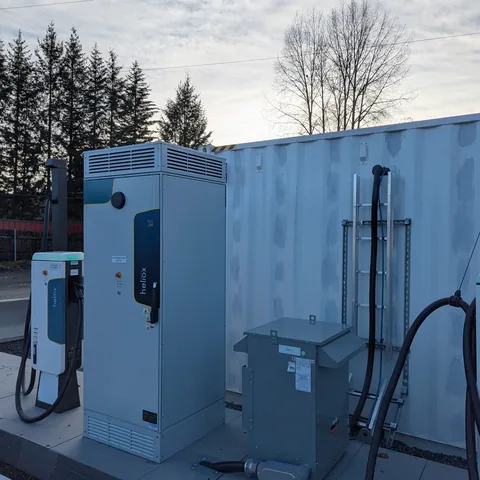 An EV charging site: a charging pedestal and power cabinet on a concrete pad against a container wall.