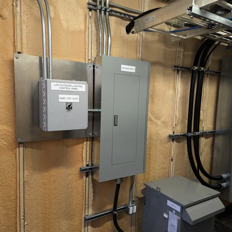 An outdoor lighting control panel beside a panelboard and transformer, with conduit run in straight lines up the wall.