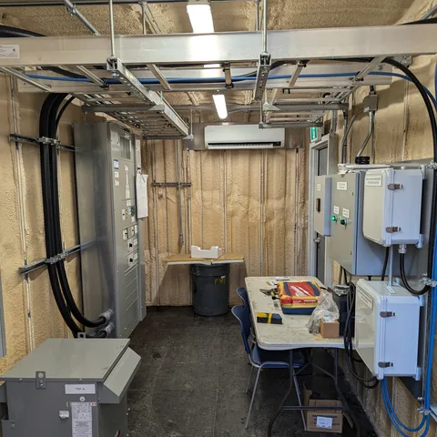 A finished service room, lights on, with panels and enclosures mounted down one wall and cable run overhead.