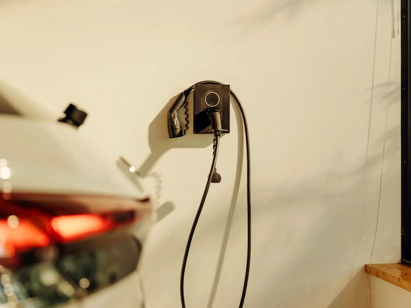An electric car plugged into a wall-mounted home charger, its cable running down to the port.